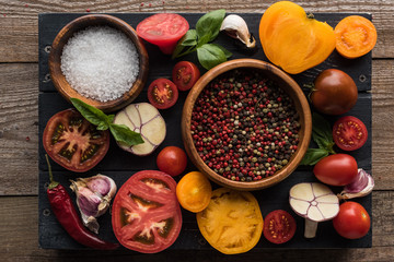 top view of bowls with pepper and salt near chilli pepper, spinach, sliced tomatoes and garlic on black tray on wooden table