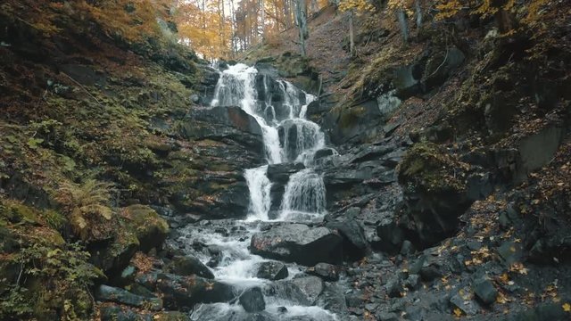 pictorial waterfall flows over large grey rocks among ground with fallen leaves on autumn day upper view. Shipot waterfall, Carpathian mountains. 4K