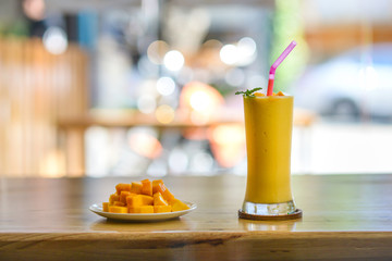 Mango smoothie in tall glass with plate of diced mangoes on wooden table.