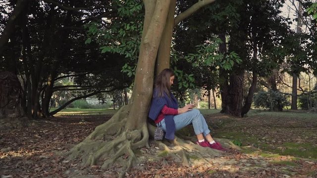 Girl in a blue sweater with a smartphone is sitting under a tree with mighty winding roots