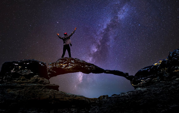 A young man on a stone bridge with the Milky Way galaxy on the mountains. Long exposure photograph, with grain.Image contain certain grain or noise and soft focus.