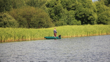 Fototapeta na wymiar Summer water landscape, a lone fisherman on a small green boat with outboard motor stands with a fishing rod at the grassy river Bank