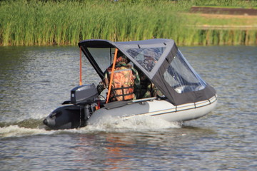 Sailor in camouflage lifejacket floating on awning PVC inflatable boat with two stroke 8 hp outboard motor on river water on hreen shore on summer day