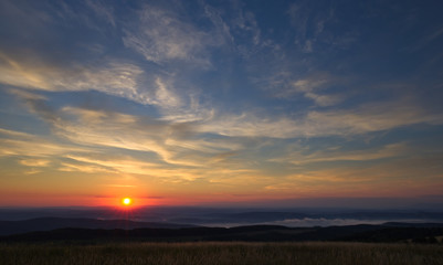 Sun right after rising above the horizon with brightly colored sky slightly cloudy above foggy hills