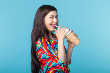 Funny portrait of pretty hipster girl wearing trendy bright clothes accessories and make up. She is drinking smoothie over blue background