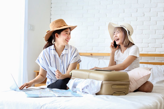 Two women are preparing to travel. Happy young friends packing clothes put in suitcase on bedroom. Female using computer laptop planning travel information. Asian people. - Powered by Adobe