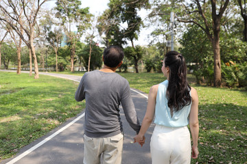 rear view middle adult asian couple walking and talking in the park with nature green trees