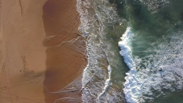 Top down aerial view of wave coming towards the sandy empty beach coastline.