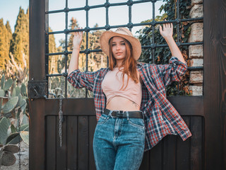 Young red-haired woman on a ranch in a straw hat, a plaid shirt and jeans on the background of the grill. Casual style