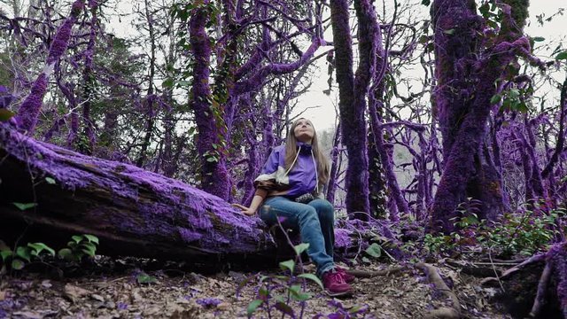 Violet fairytale forest. Happy girl in the casual style is sitting on the moss-covered purple tree and looks up in a boxwood forest. Fantasy, unreal, fairytale atmosphere
