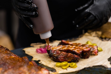 Grill restaurant kitchen. Chef in black cooking gloves pouring sauce over sliced smoked pork ribs.