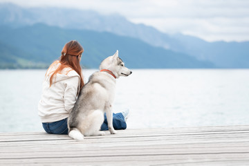 A young woman with brown hair and white sweater is sitting at the pier at the lake with calm water. A Siberian husky female dog is lying down near the girl. The Alps mountains in the background.