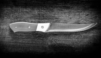 Black and white photo of a kitchen knife, with yellow and white handle and steel sawtooth blade on a natural wood background.