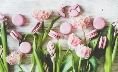 Flat-lay of sweet pink macaron cookies and fresh spring flowers over white wooden background, top view. Food texture, background and wallpaper