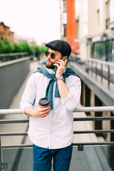 Young man with beard in sunglasses holding coffee cup and talking on mobile phone while walking outdoors. Always in touch.