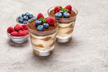 Classic tiramisu dessert with blueberries and raspberries in a glass and bowls with berries on concrete background