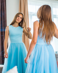 Fototapeta premium portrait of young teenager brunette girl with long hair in blue dress looking in the mirror