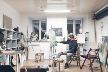 Professional artist sits on a chair in the interior of a cozy artistic studio and is working on a...