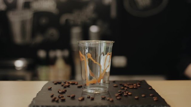 Male barista putting an empty clear plastic cup with some caramel inside on the bar counter with coffee beans. A close view