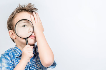 Cute boy looks into a magnifying glass with one eye, the other covered with his hand on a white...