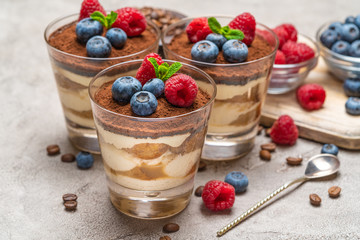 Classic tiramisu dessert with blueberries and raspberries in a glass and bowls with berries on concrete background