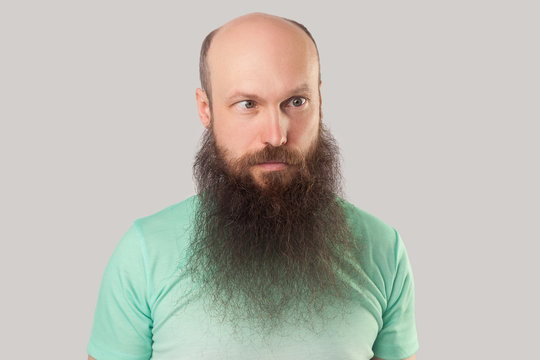 Portrait of crazy middle aged bald man with long beard in light green t-shirt standing and looking with croosed eyes funny face. indoor studio shot, isolated on grey background.