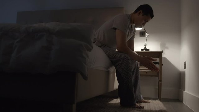 Young man sitting on the edge of the bed at night rubbing his hands suffering with anxiety, stress and depression.