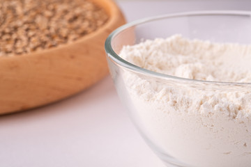 Wheat seeds grains in wooden bowl near with wheat flour in glass bowl, angle view, isolated on white background, copy space