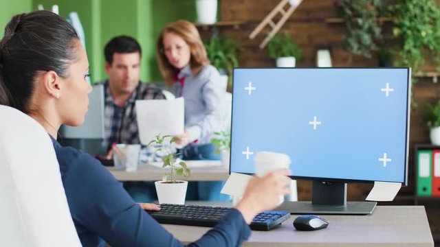 Young manager female working on her office computer with an isolated blue screen. Mock-up display. Coworkers in the background.