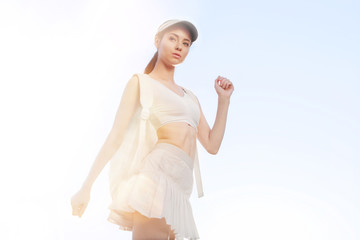 Fototapeta na wymiar Sports woman posing in fashion sportswear on sunny sky background. Beautiful slim girl in trendy white skirt, top, cap and backpack. 