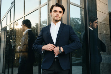 Young handsome bearded brunette man in white shirt and classic jacket standing dreamily looking in camera with glass building on background