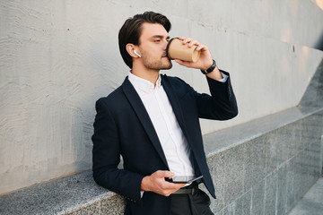 Young handsome bearded brunette man in white shirt and classic suit with wireless earphones holding cellphone in hand dreamily drinking coffee while standing near wall on street