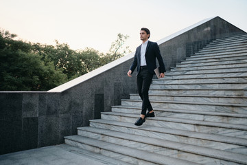 Young handsome brunette man in white shirt and classic suit going down the stairs on street with laptop in hand and dreamily looking aside alone