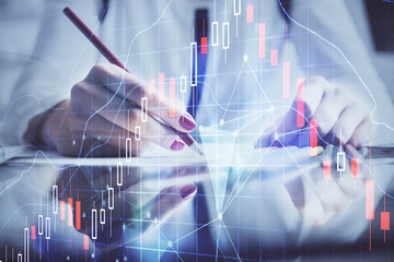 A woman hands writing information about stock market in notepad. Forex chart holograms in front. Concept of research.