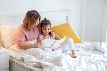 Happy little cute asian girl and her senior retired grandmother lying on the bed. Grandma smile and teach her preschool grandchild do homework or write on the book on the bed in bedroom.