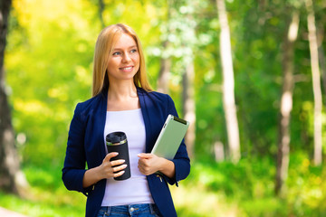 Young happy smiling caucasian adult girl business student walking concept in the summer green park outdoor holding tablet and coffee thinking of credit money finance people job in the college