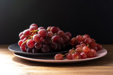 Bunches of ripe purple grapes on plates on a wooden table on a black background in the sun. Side view. 