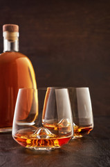 Two crystal glasses of whiskey and a full bottle on a wooden table.