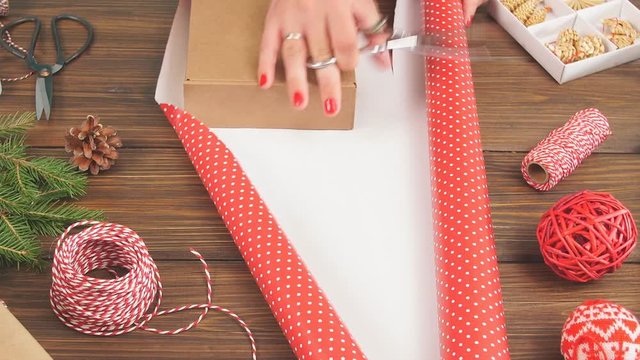 Woman wrapping gift box with equipment and decorating items on wood table, preparing for celebrating Christmas holidays.