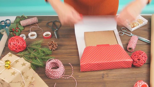 Female hands wrapping Christmas gift in craft ecological paper on dark wooden background with decorating elements, colourfil twine stripes and pack paper rolls.