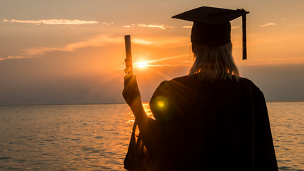 Rear view of a college graduate with a diploma in his hand on a sunset background over the sea. New...