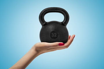 Side closeup of woman's hand facing up and holding big black 32 kg kettlebell on light blue...