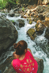 Image of a young Caucasian woman, wearing red shirt, from back while she is sitting on a big rock and watching the flowing water of a waterfall and river. Big rocks and trees surrounding the cascade.