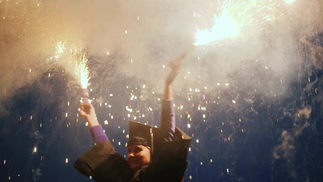 A joyful graduate in a mantle and cap waving two fireworks. Graduation party