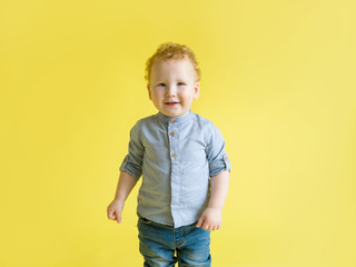 Red-haired curly baby in a blue shirt smiling looking at the camera on a yellow background