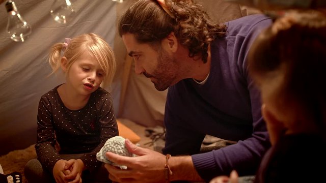 Dad with daughters plays with a toy whale in a tent at home.
