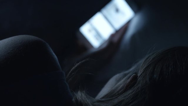 A woman scrolling on her mobile phone in bed at night. The bright blue screen light illuminating her face.