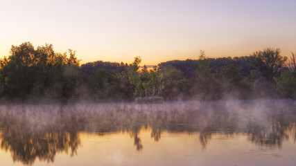 Panorama of tendrils of morning mist on a lake at sunrise and a colorful orange glow in the sky reflected in the tranquil water with the surrounding woodland trees in an atmospheric landscape
