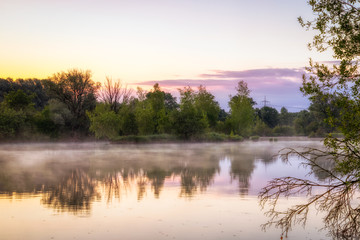 Fototapeta na wymiar Panorama of tendrils of morning mist on a lake at sunrise and a colorful orange glow in the sky reflected in the tranquil water with the surrounding woodland trees in an atmospheric landscape