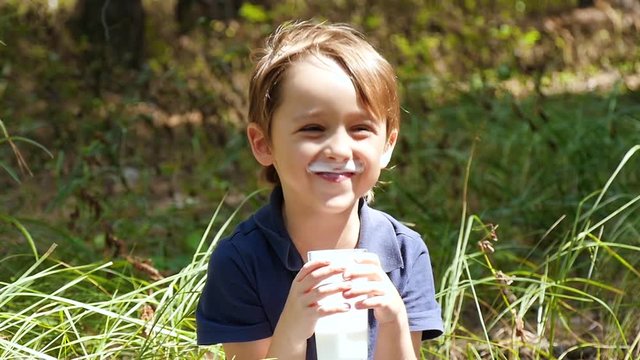 A happy child in nature loves to drink kefir from a dairy product from a transparent glass cup in slow motion